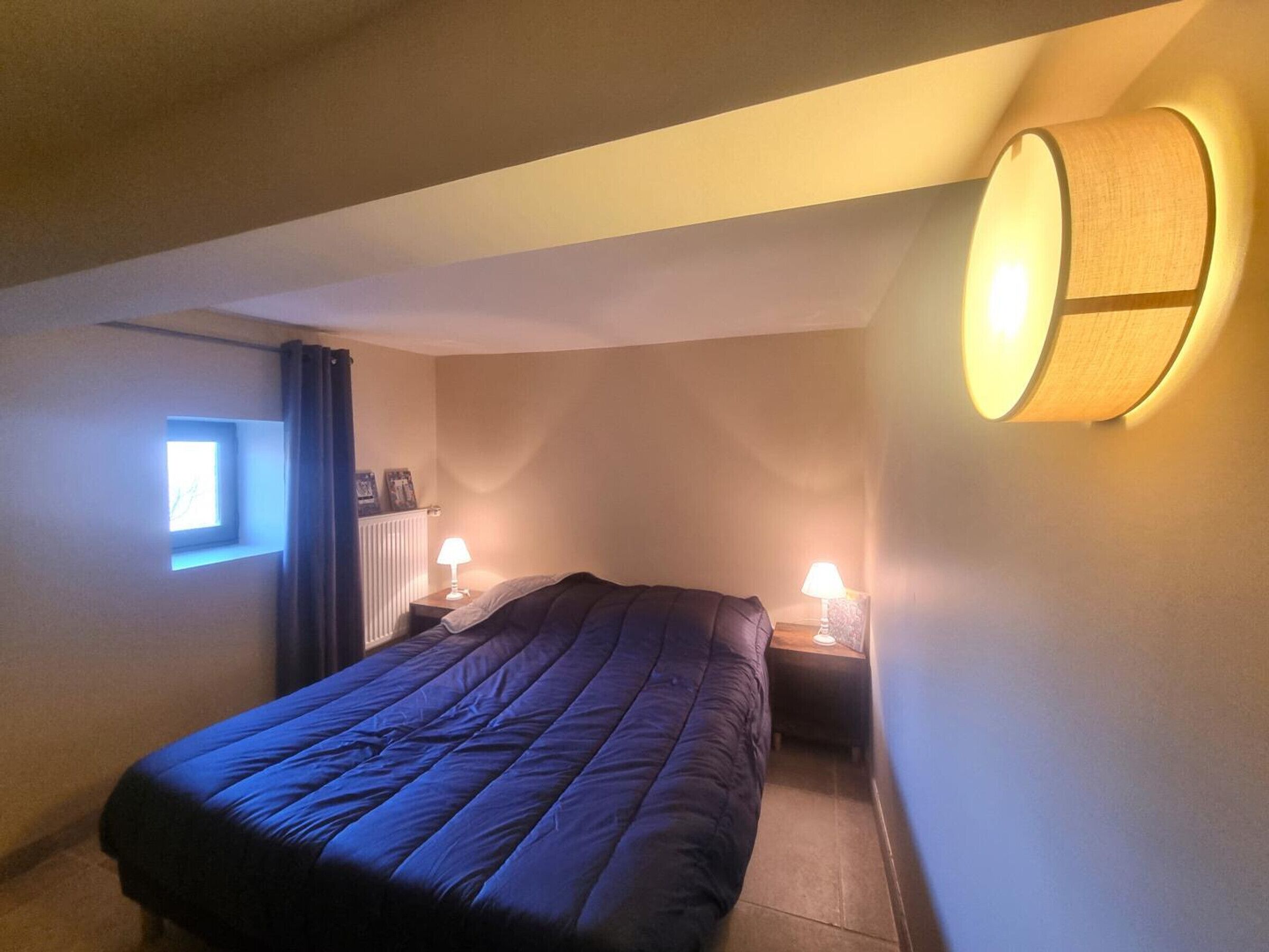 A small, dimly lit room with a single bed, two bedside lamps, and a window with curtains.