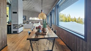A modern interior with a wooden dining table, a hanging light fixture, and a large window offering a view of a snowy landscape.