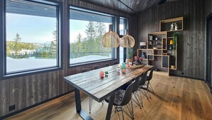 A dining area with a wooden table and chairs, large windows with a view of trees and snow, and a bookshelf with various items.