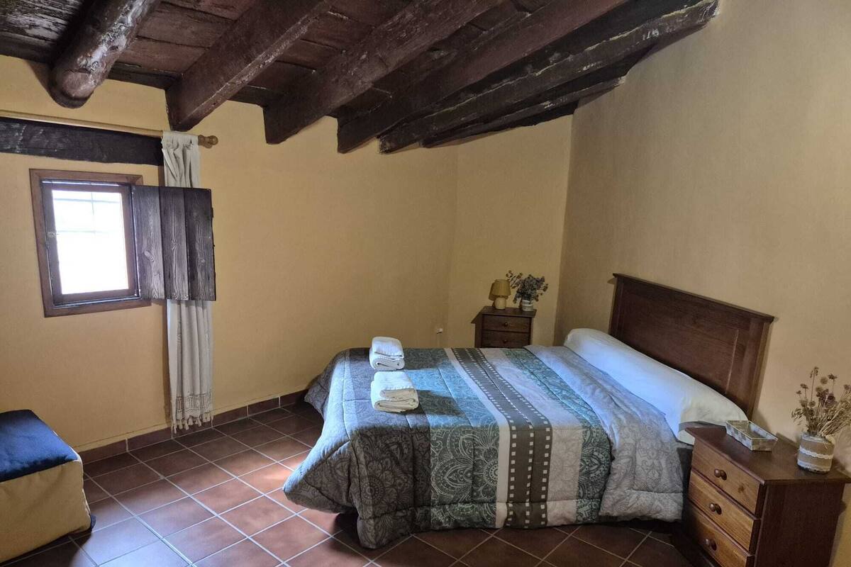 A bedroom with a bed, a nightstand, a dresser, a window with curtains, and a wooden ceiling.