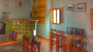 A dining area with wooden tables and chairs, a tiled backsplash, and a kitchen area with a wooden counter and hanging lights. - Cau de l'Art (Llucena)