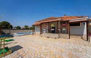 Una casa con un patio de piedra, una piscina y un porche cubierto adornado con cestas de flores colgantes.