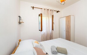 Un dormitorio con cama, armario, ventana con cortinas y una estantería de pared con una figurita de gato.
