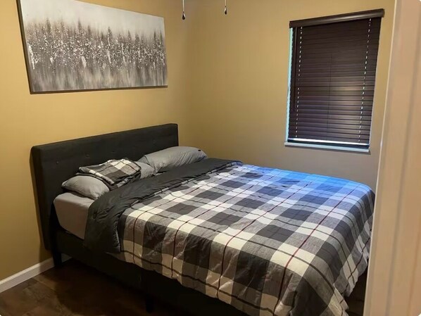 Un dormitorio con cama, ventana con persianas, un cuadro en la pared y una puerta.