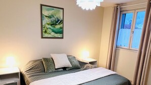 A bedroom with a bed, two bedside tables, a wall-mounted lamp, a ceiling light fixture, a window with curtains, and a painting on the wall. - Apartment in the heart of the High Vosges! (Cornimont)