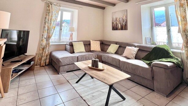 A modern living room with a sectional sofa, a wooden coffee table, and a television on a wooden stand. There are windows with curtains, a painting on the wall, and a tiled floor. - Apartment in the heart of the High Vosges! (Cornimont)