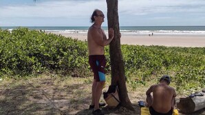 A beach scene with a man standing under a tree, another man sitting on a mat, and a beach in the background.