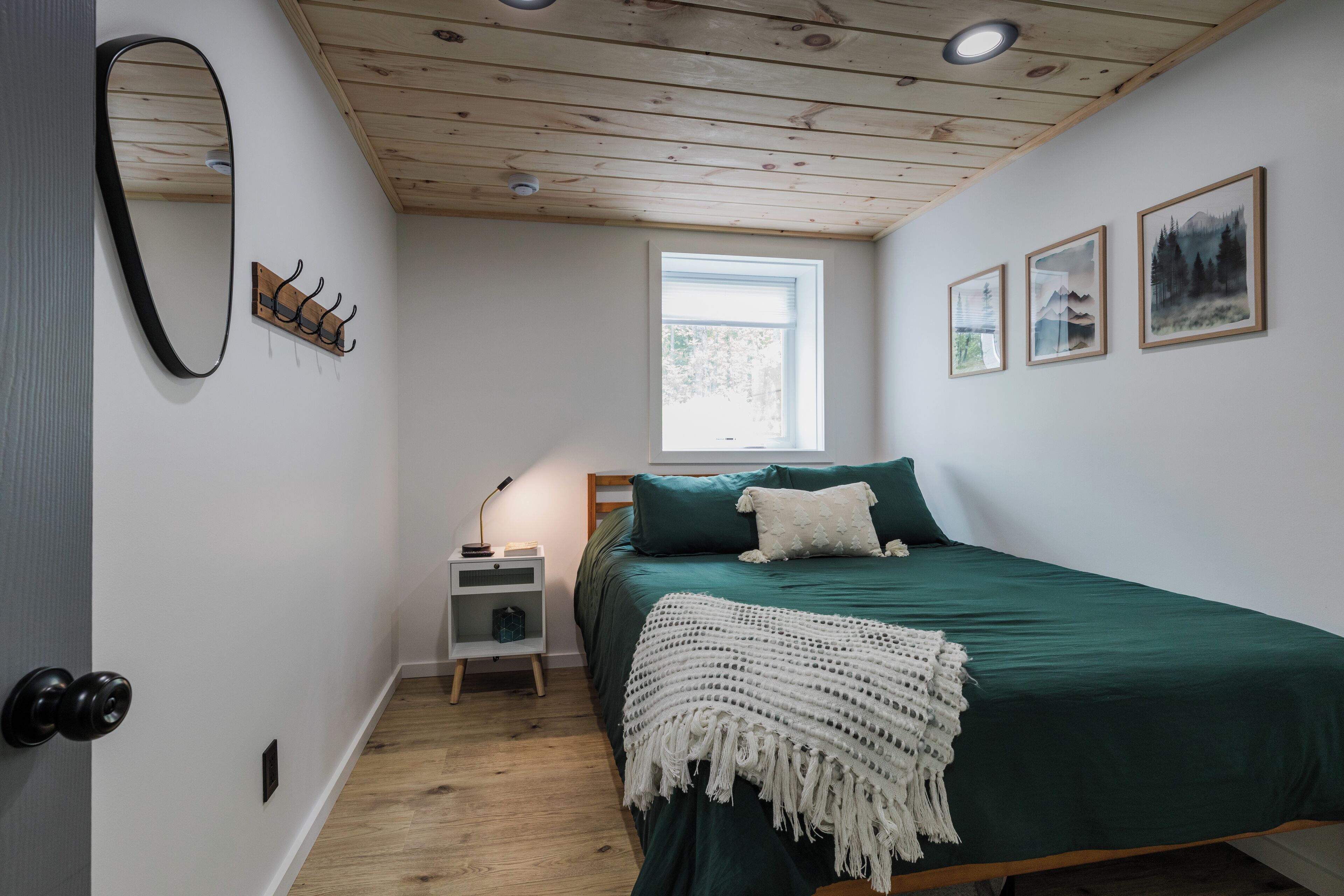 Un dormitorio con techo de madera, una cama con ropa de cama verde, un edredón blanco, una mesita de noche con lámpara, un espejo y tres cuadros en la pared.