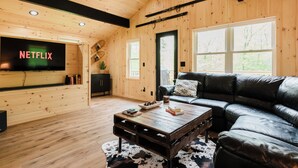 A cozy living room with a wooden interior, a black leather sofa, a coffee table, and a TV mounted on the wall. - The Mont Haus- WOOD FIRED SAUNA - Minutes to Mt. Snow and Stratton! (Wardsboro)
