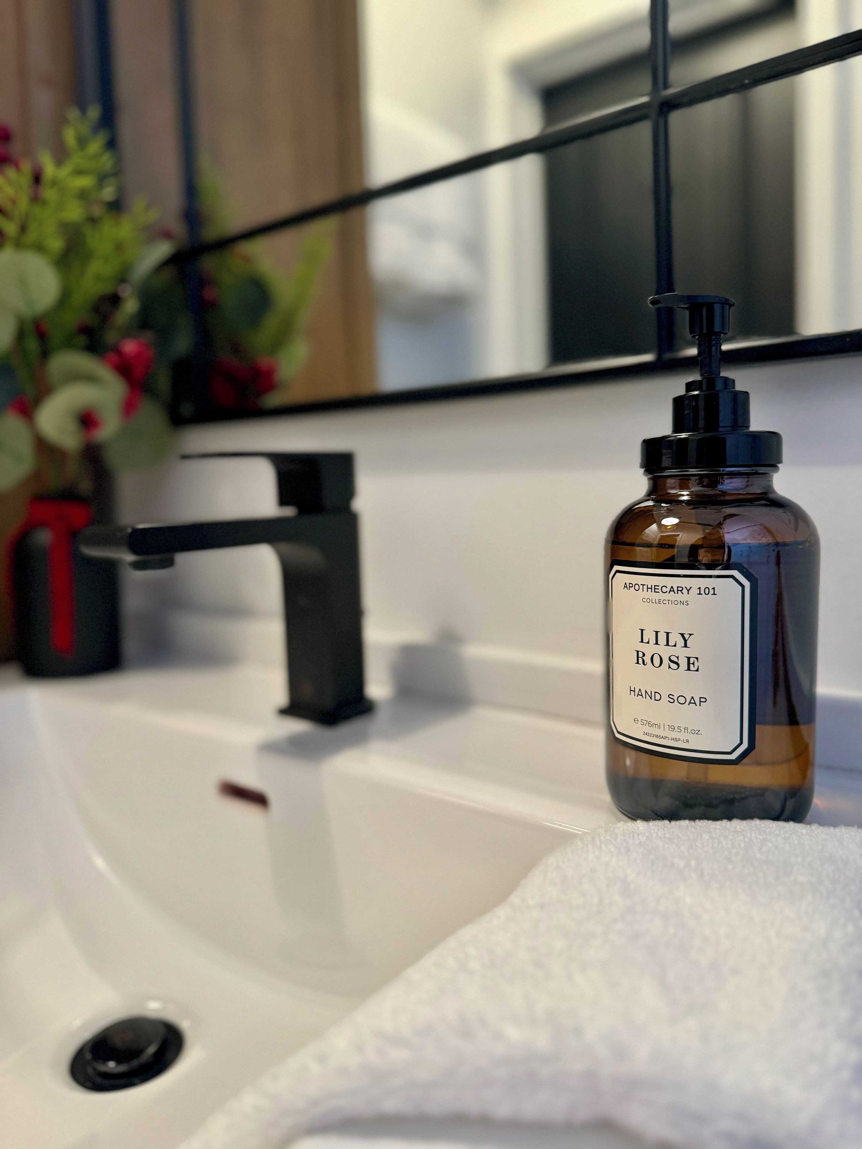 Un lavabo de salle de bain avec un flacon de savon liquide étiqueté « LILY ROSE » et un miroir au-dessus.