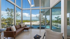 A modern living room with a large glass wall, a brown leather sofa, and a view of a pool and beach.