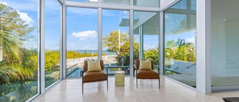A modern interior with large glass windows, two brown leather chairs, a small round table, and a view of a beach and palm trees.
