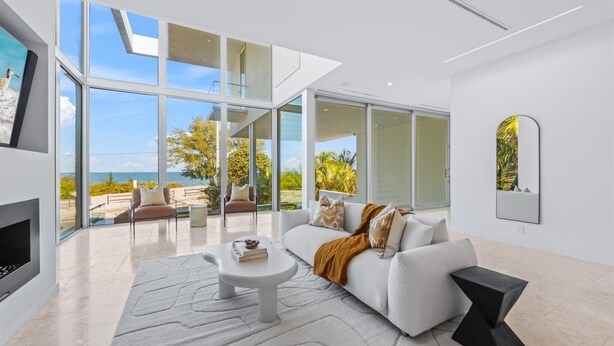 A modern living room with a white sofa, a coffee table, and a fireplace. There is a large window offering a view of the outdoors.