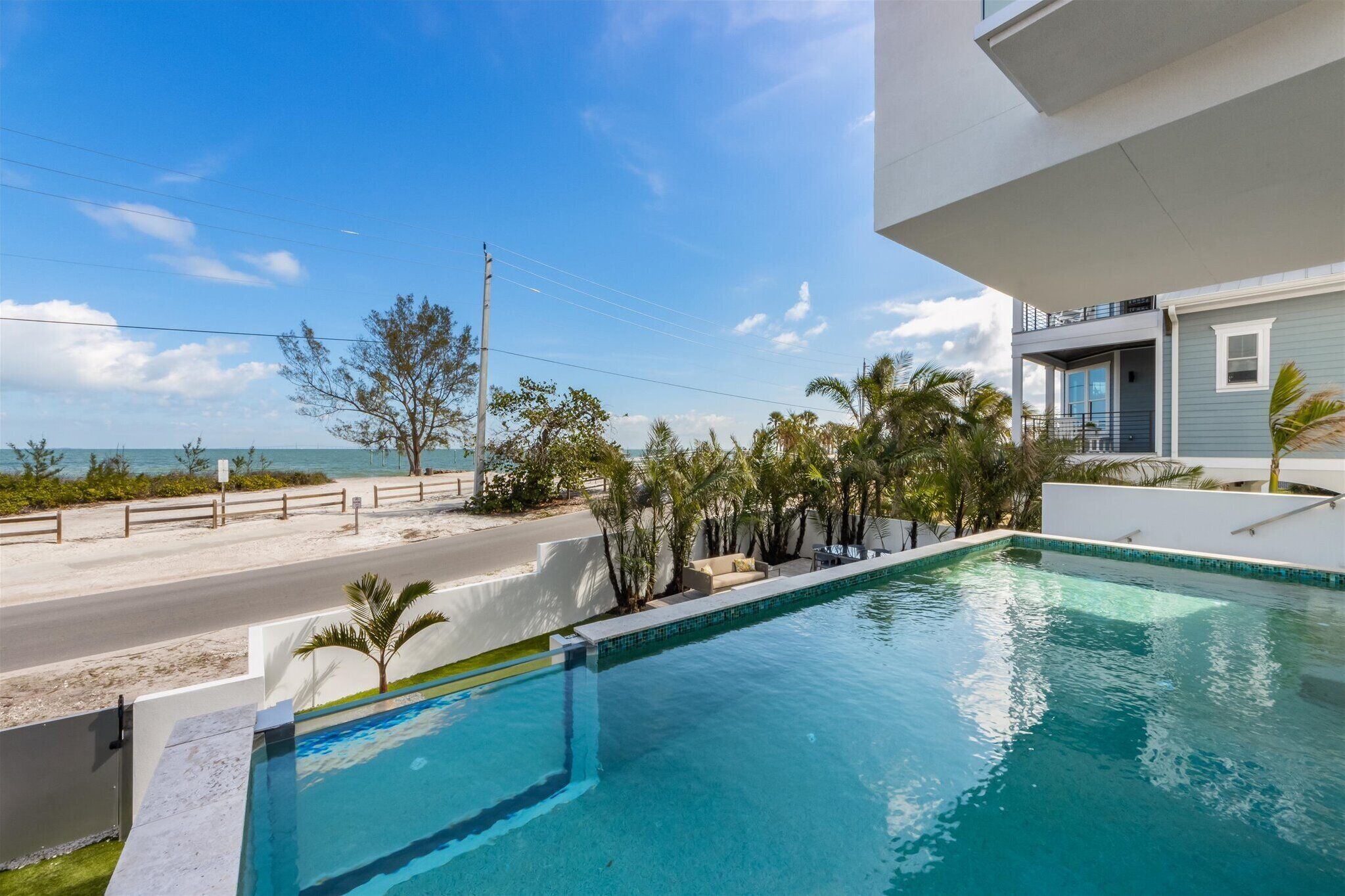 A pool with a clear view of the beach and ocean, surrounded by palm trees and a modern building.