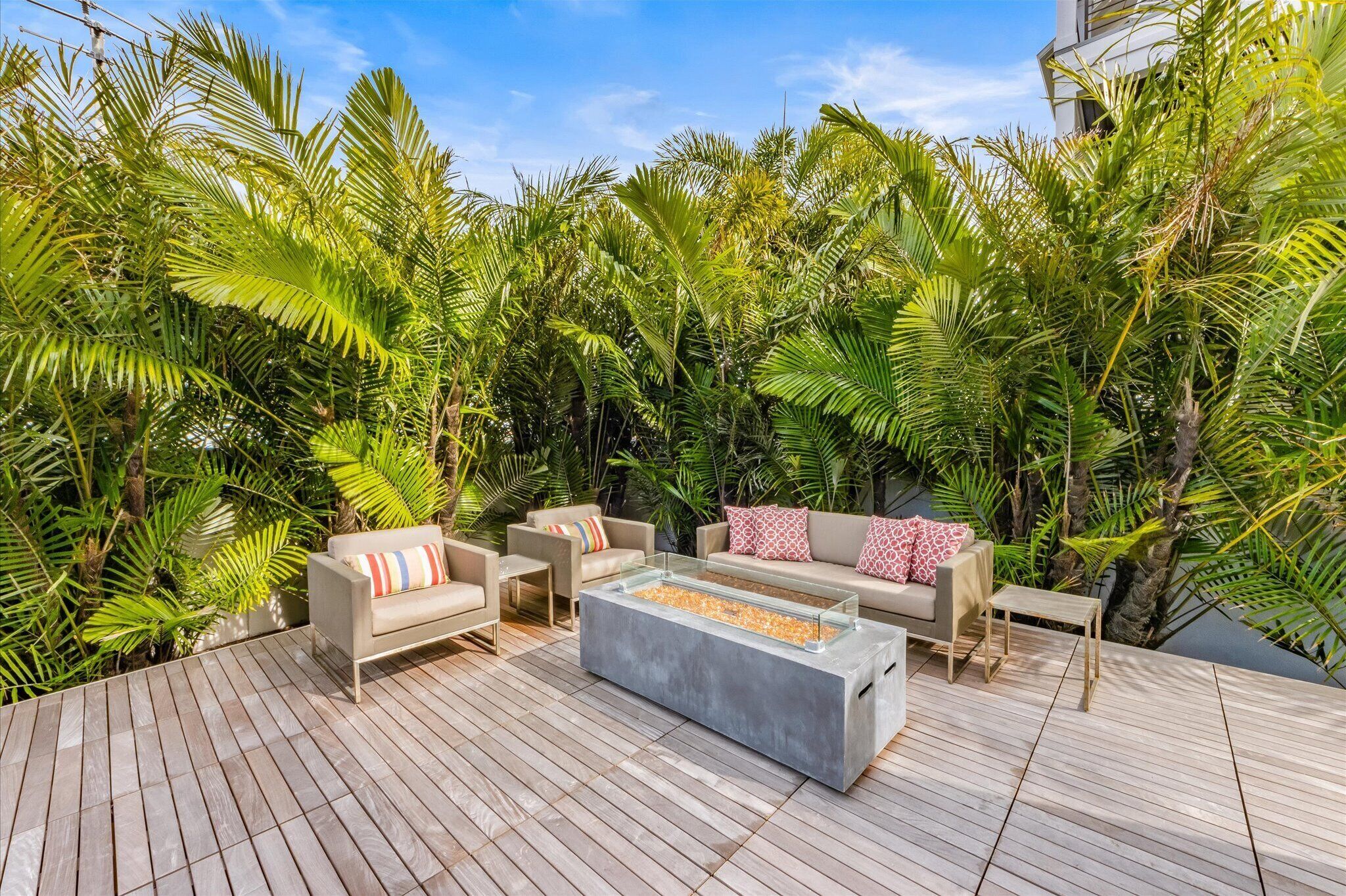 A modern outdoor seating area with a glass-top coffee table, cushioned sofas, and a wooden deck surrounded by lush tropical foliage.