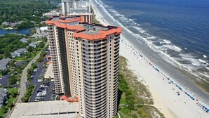 Un hôtel de grande hauteur à la façade incurvée, situé à côté d’une plage parsemée de parasols et d’un parking.