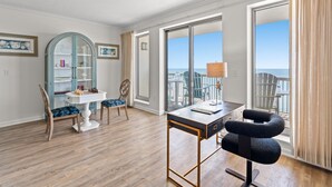 Une chambre d’hôtel moderne avec un plancher en bois, un coin repas comprenant des chaises et une table, une grande fenêtre donnant sur la mer et un balcon agrémenté de rideaux.
