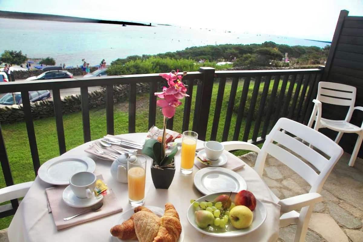 Ein Frühstückstisch mit Croissants, Obst und Saft, mit Blick auf eine schöne Landschaft aus Meer und geparkten Autos.