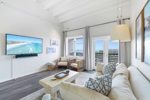 A living room with a large flat-screen TV, a sofa, a coffee table, and a view of the sea through the balcony doors.
