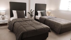 A hotel room with two beds, each with a dark headboard and a bedside table with a lamp. - INAS 2 BEDROOM APARTMENTS (Corfu)