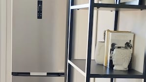 A modern kitchen with a stainless steel refrigerator, a black metal shelving unit, and a white wall with a framed picture and decorative plants. - INAS 2 BEDROOM APARTMENTS (Corfu)
