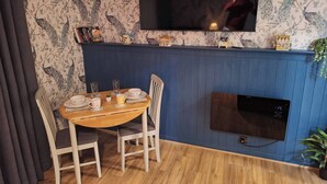 A dining area with a wooden table, white chairs, a blue wall, a mounted TV, and a small shelf with decorative items. - Ouston Oasis, Beamish (Chester-le-Street)