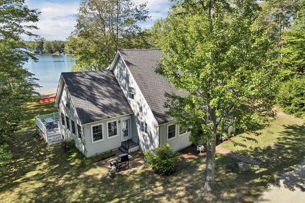 A two-story house with a sloped roof, a front porch, and a small deck. - Lake Winnipesaukee Waterfront Home Private Beach Dock Sleeps 10 Sunset Views (Moultonborough)