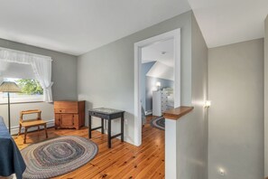 A room with a wooden floor, a small wooden chair, a cabinet, a table, and a doorway leading to another room with a lamp and a dresser. - Lake Winnipesaukee Waterfront Home Private Beach Dock Sleeps 10 Sunset Views (Moultonborough)