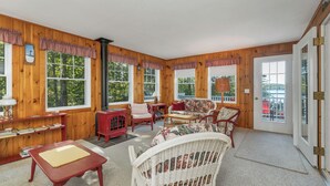 A cozy room with wooden walls, large windows, a sofa, chairs, a table, and a lamp. - Lake Winnipesaukee Waterfront Home Private Beach Dock Sleeps 10 Sunset Views (Moultonborough)