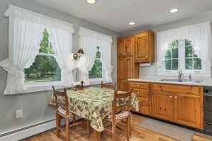 A kitchen with wooden cabinets, a dining table with chairs, and large windows with white curtains. - Lake Winnipesaukee Waterfront Home Private Beach Dock Sleeps 10 Sunset Views (Moultonborough)