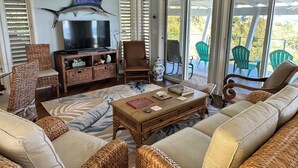 A living room with wicker furniture, a television, and a large window with a view of outdoor chairs. - Diamond in Paradise-Hear the waves, feel the breezes from this 3 bedroom villa! (Christiansted)