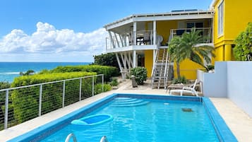 A swimming pool with a clear view of the ocean, a covered outdoor area with a staircase, and a yellow building with balconies.
