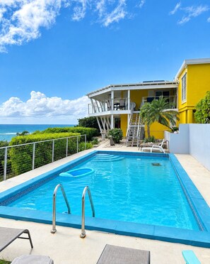 A swimming pool with a clear view of the ocean, a covered outdoor area with a staircase, and a yellow building with balconies. - Diamond in Paradise-Hear the waves, feel the breezes from this 3 bedroom villa! (Christiansted)