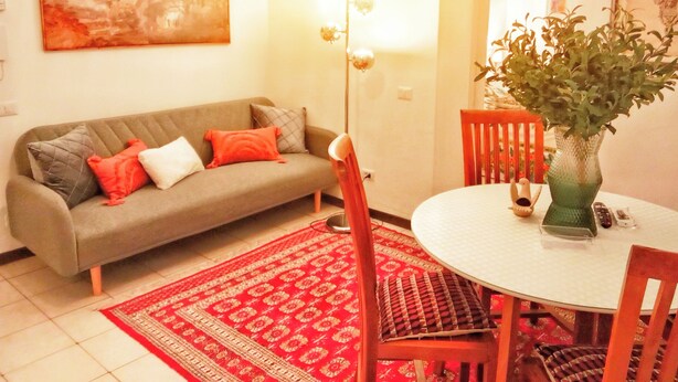 En stue med en sofa, et spisebord med stole, en rød måtte med et mønster, og en grøn vase med en plante.