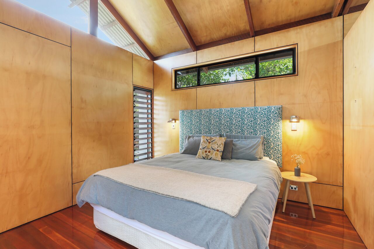 A bedroom with a large bed, wooden walls, a window with blinds, and a small bedside table with a vase.