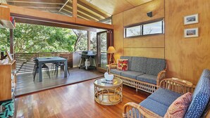 Amplia sala de estar con suelo de madera, un sofá, una mesa de centro y una mesa de comedor con sillas. Un gran ventanal ofrece vistas al exterior.