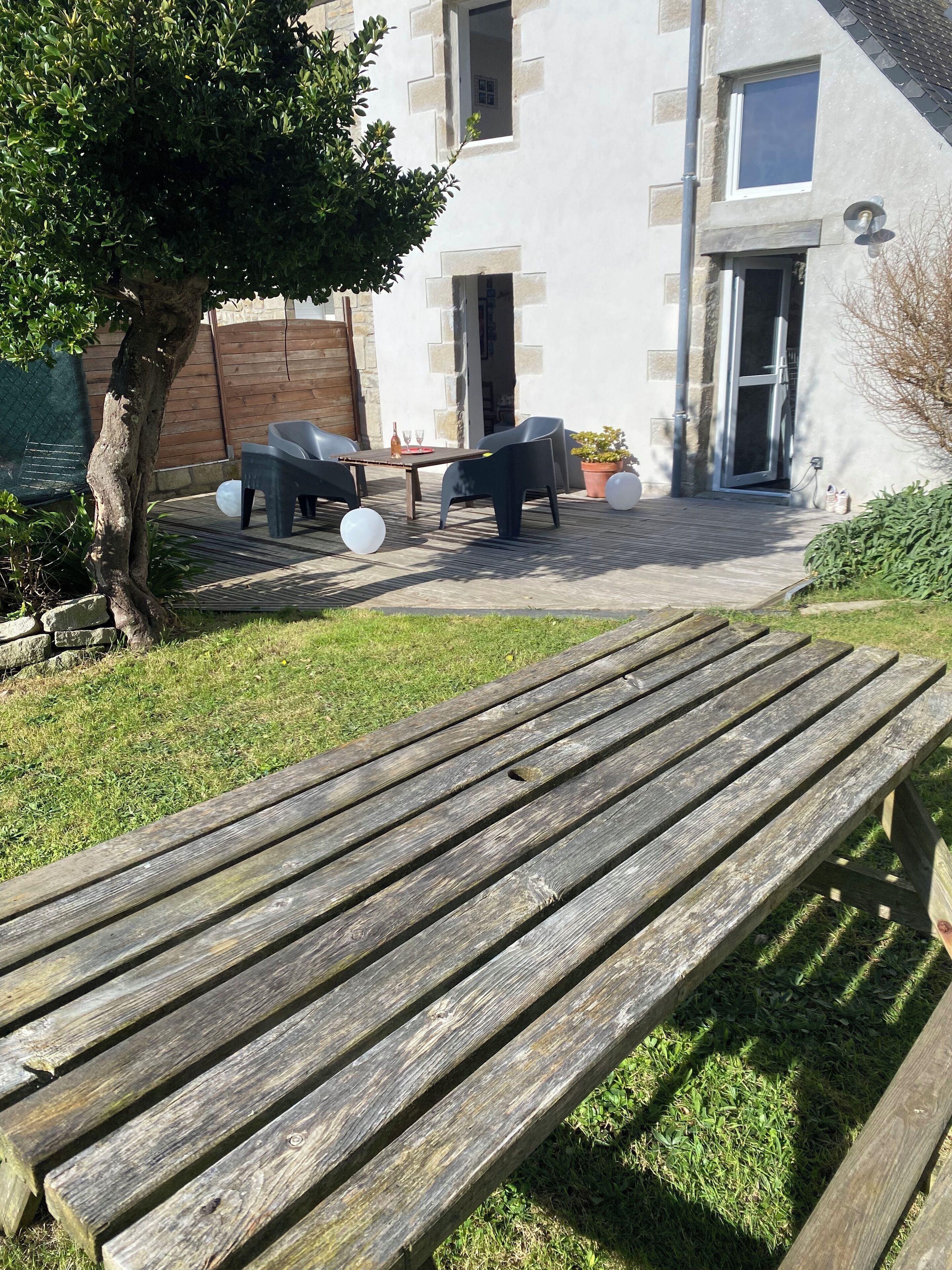 A wooden bench in a garden area with a patio table and chairs, a tree, and a building in the background.