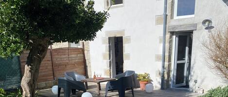 A wooden bench in a garden area with a patio table and chairs, a tree, and a building in the background.