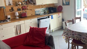 A cozy kitchen with wooden cabinets, a dining table with a colorful tablecloth, and a red sofa. - Charming fisherman's cottage (Île-de-Sein)