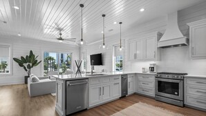 A modern kitchen with white cabinetry, a central island, stainless steel appliances, pendant lighting, and a large window offering a view of palm trees.