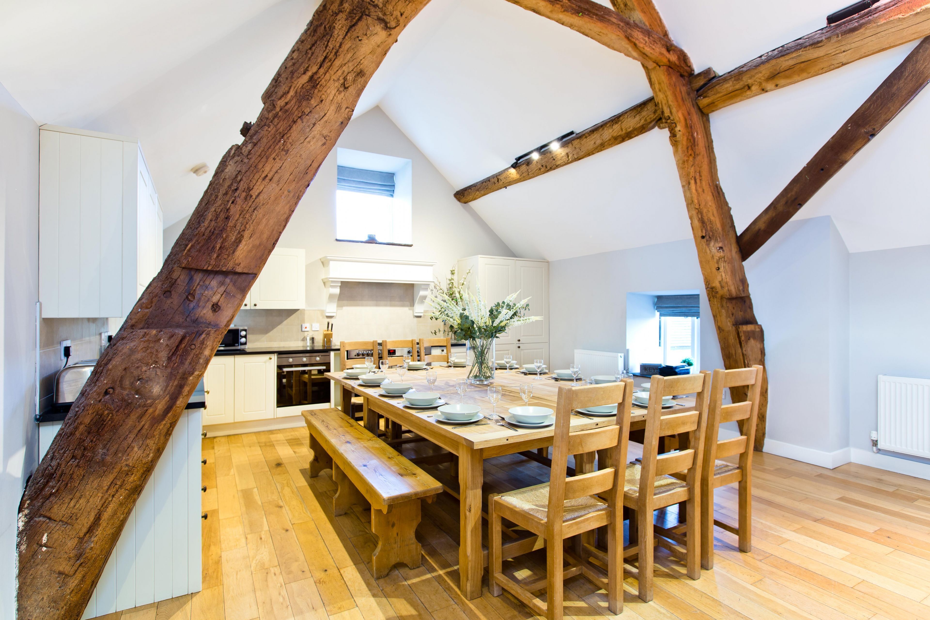 A spacious kitchen with wooden beams, a dining table set for six, and a large window providing natural light.
