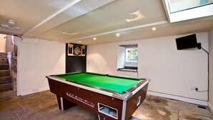 Une table de billard dans une pièce dont le sol est recouvert de pierres, avec un jeu de fléchettes, une petite fenêtre et un téléviseur fixé au mur.