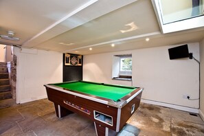 A pool table in a room with a stone floor, a dartboard, a small window, and a television mounted on the wall.
