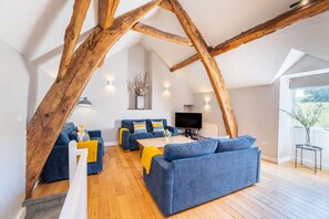 Un loft spacieux avec des poutres apparentes, un salon doté de canapés bleus et de touches de couleur jaune, un téléviseur à écran plat et une grande fenêtre offrant une vue sur la verdure.