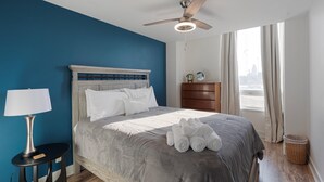Ein Hotelzimmer mit Bett, Nachttisch, Lampe, Kommode und Blick auf die Stadt durch das Fenster.
