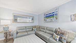 A living room with a sectional sofa, a side table with a lamp, and a painting of a seaside scene. - Happy OURS 206–Pool Near Beach–Sleeps 6 (Panama City Beach)