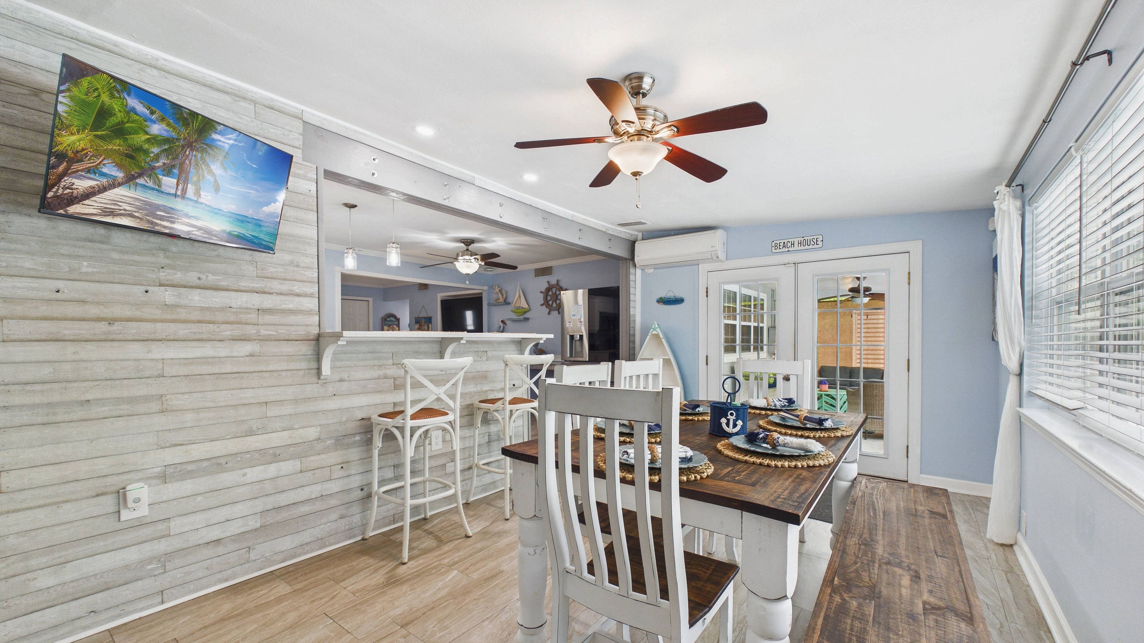 Un espace repas décoré sur le thème de la plage, avec des chaises blanches, une table en bois et un ventilateur de plafond.