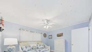 Une chambre à coucher comprenant un lit, une table de chevet, une lampe, un ventilateur de plafond, une fenêtre avec des stores, une porte et des éléments de décoration murale, notamment un gouvernail de navire et un tableau.