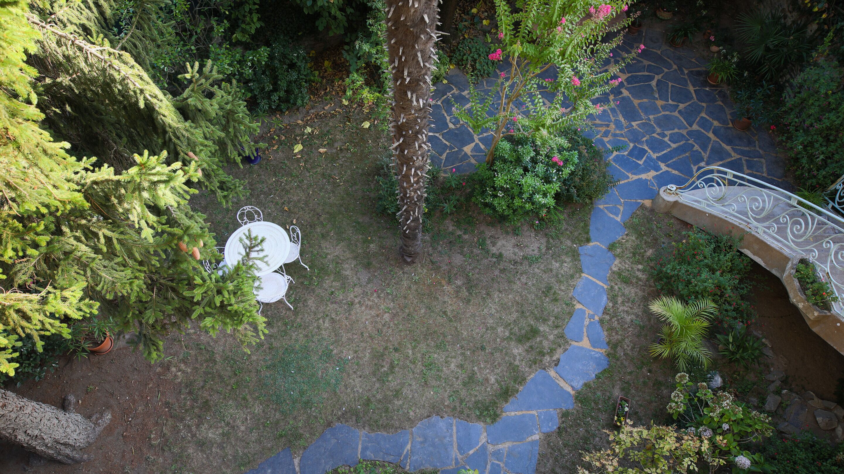 Een tuin met een pad van stenen, een witte metalen tafel en stoelen, en diverse planten.