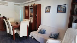 A living room with a white sofa, two decorative pillows, a wooden cabinet, and a dining area with a table and chairs. - Oasis  1º Solo Familias! (Xeraco Playa)